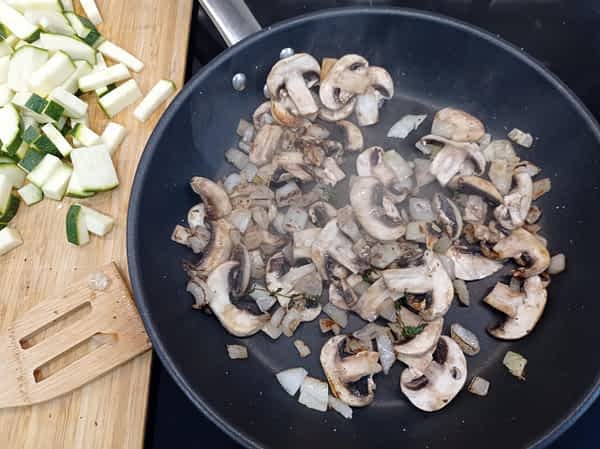 cebolla y champiñones en un sartén con trozos de calabacín al lado