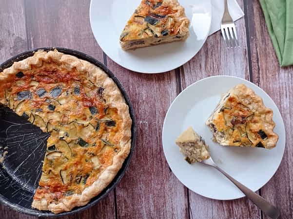 dos trozos de quiche de calabacín y champiñones en platos al lado del quiche entero